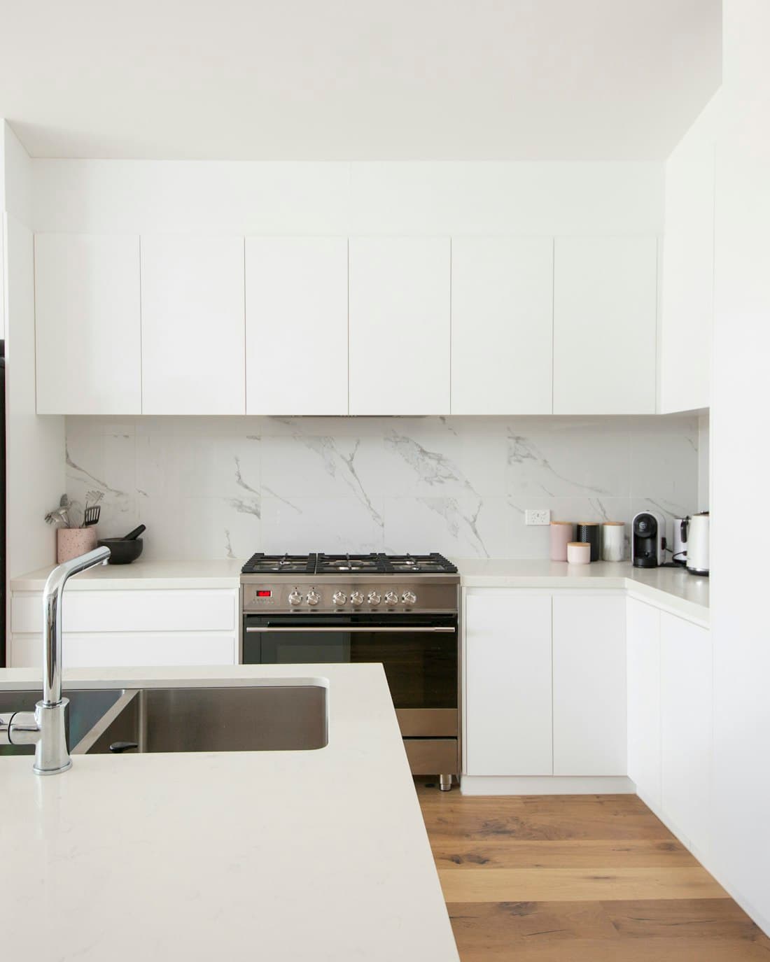 A clean white kitchen with stone worktops, a stainless sink and natural daylight.